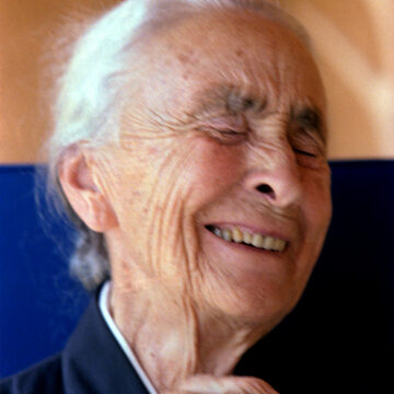 Photograph of Georgia O'Keeffe laughing with her eyes closed. Her white hair is tied up. and her hand rests by the collar of her black dress.