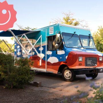 Photograph of the Art to G.O. truck with colorful cloud motifs on it and its awning near some bushes and trees. In the top left corner of the image is a sticker of a pink smiley face, the Risolana logo.