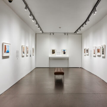 A view of a gallery with white walls and framed color photographs. The room has a dark brown floor with a leather bench in the center.