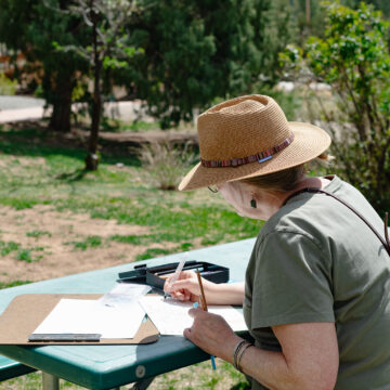 A person with a hat sits at a table sketching and painting. Behind them are green trees and grass.