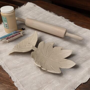 Photograph of two large leaves bent into bowls made of clay placed on a piece of fabric on a wooden table. Around the clay leaf bowls are tools for clay sculpting such as a rolling pin and sponge.