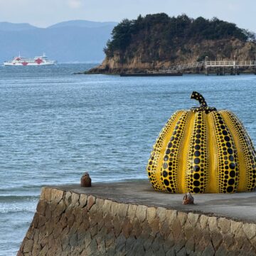 Photograph of a sculpture of a yellow pumpkin with black dots sitting on a concrete pier near the ocean. Behind the pumpkin, water extends with a small island on the horizon and a boat can be seen in the background.