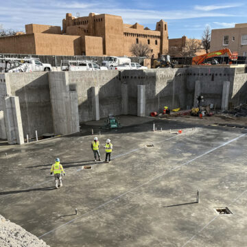 Photograph of a large construction site, where a concrete foundation and walls have been built. At the center of the site a few workpeople with hard hats and bright vests stand. Above the site an adobe building is visible in the background.