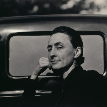 Quarter length portrait of O'Keeffe dressed in black with her hair pulled back leaning against a car. She looks at the camera with her face in semi-profile wearing a slight smile with her hand leaning against her cheek.