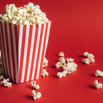 A pin-stripped container holding popcorn against a red background. Some popcorn pieces are also strewn about.