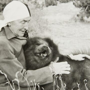 Black and white photograph of Georgia O'Keeffe wearing a jacket and a scarf wrapped around her head. O'Keeffe sits on the ground embracing a Chow Chow dog.