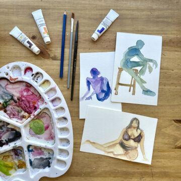 Three small watercolor paintings of figures in shades of blue, purple, and beige. Nearby on the same table rests a colorful watercolor palette and a few paint tubes and brushes.