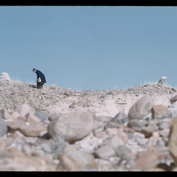 Photograph of two people very small and bent over looking at rocks against a blue sky.