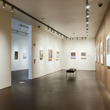 A long gallery view showing paintings on walls. Two walls are beige and the one on the right a deep tan. The entrance to the gallery is on the left. The floor is a deep gray color reflecting the walls.