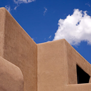 View of adobe style roof of the Georgia O'Keeffe Museum. Above the roof is bright blue sky with a cascading cloud.