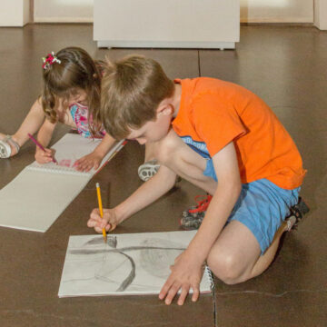 Two children are sitting on the foreground of this photograph. They are holding pencils and intently sketching abstract art.