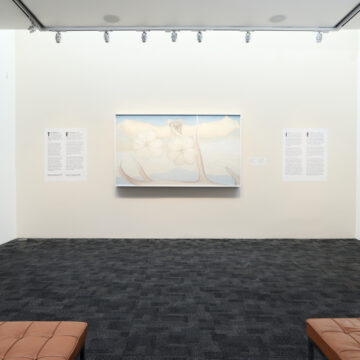 View of a gallery installation showing a painting in light pastel blues and tans against a white wall. There are two text panels on the sides. The room has a dark gray floor and two edges of leather benches are extending off the image at the bottom.