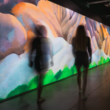 Shadows of two human figures slightly blurred as if in motion, they walk past a colorful wall that looks like a mountain landscape of with warm yellows in the background and cool blues and green in the foreground.
