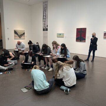 A group of students are gathered in the middle of the Georgia O'Keeffe Museum gallery. They are sitting on the floor and on benches hunched over with art supplies. Paintings are hung in the gallery.