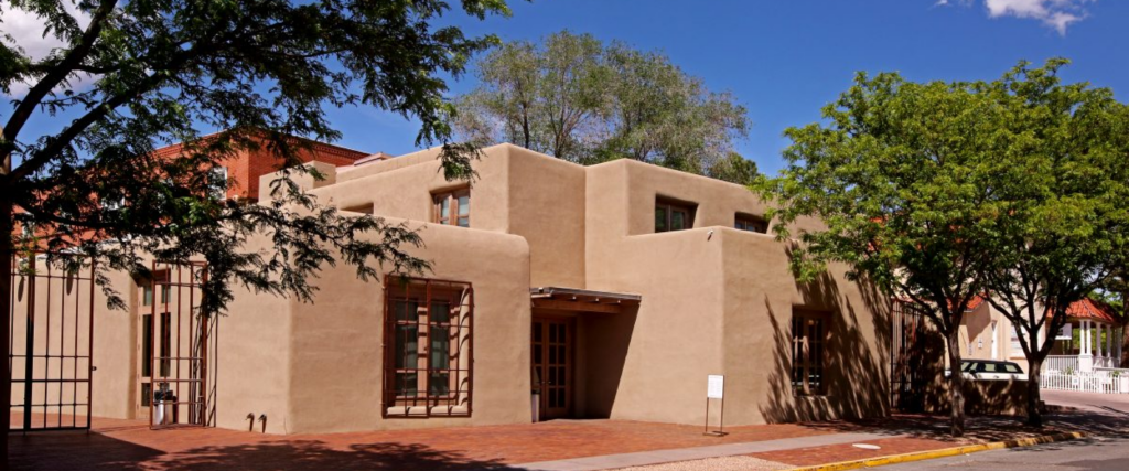 The exterior of a mud-colored stucco building in southwestern pueblo style building with a very blue sky and dramatic shadows. The building shows an entrance and a window with a gate. Three trees surround the building with a brick colored patio leading to a street.