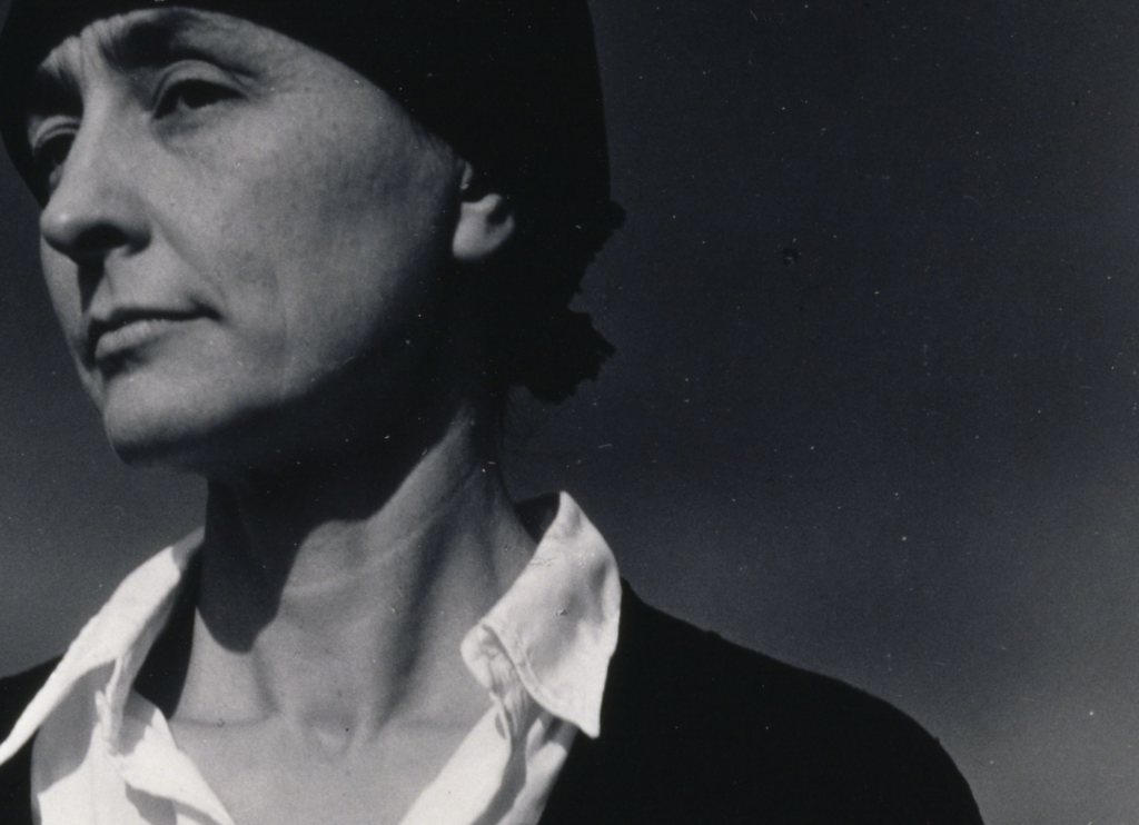 A black and white portrait of Georgia O'Keeffe. O'Keeffe wears a fabric covering over her head and is dressed simply with a white button up and black jacket.