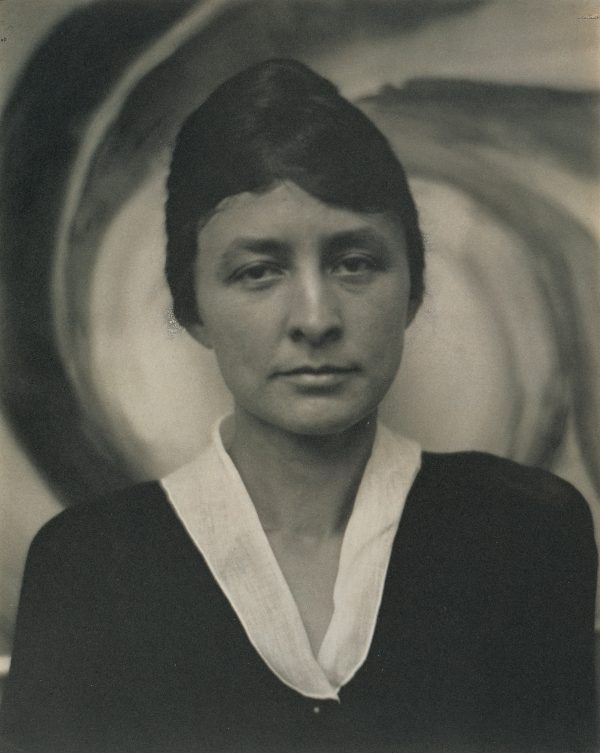 A woman stands in front of a painting with a blank expression. Her dark hair is in a bun and she wears a dark shirt with a white collar. The painting in the background is in a spiral shape. The image is in black and white.