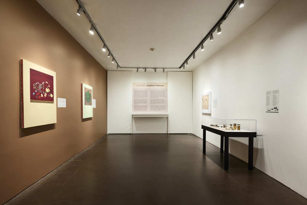 A long gallery view showing paintings on walls with a case on the left. Two walls are beige and the one on the left is a deep tan. The floor is a deep gray color reflecting the walls.