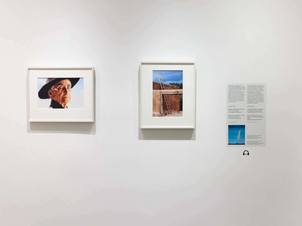 A view of a gallery with white walls and two framed color photographs and a text panel. The room has a dark brown floor.