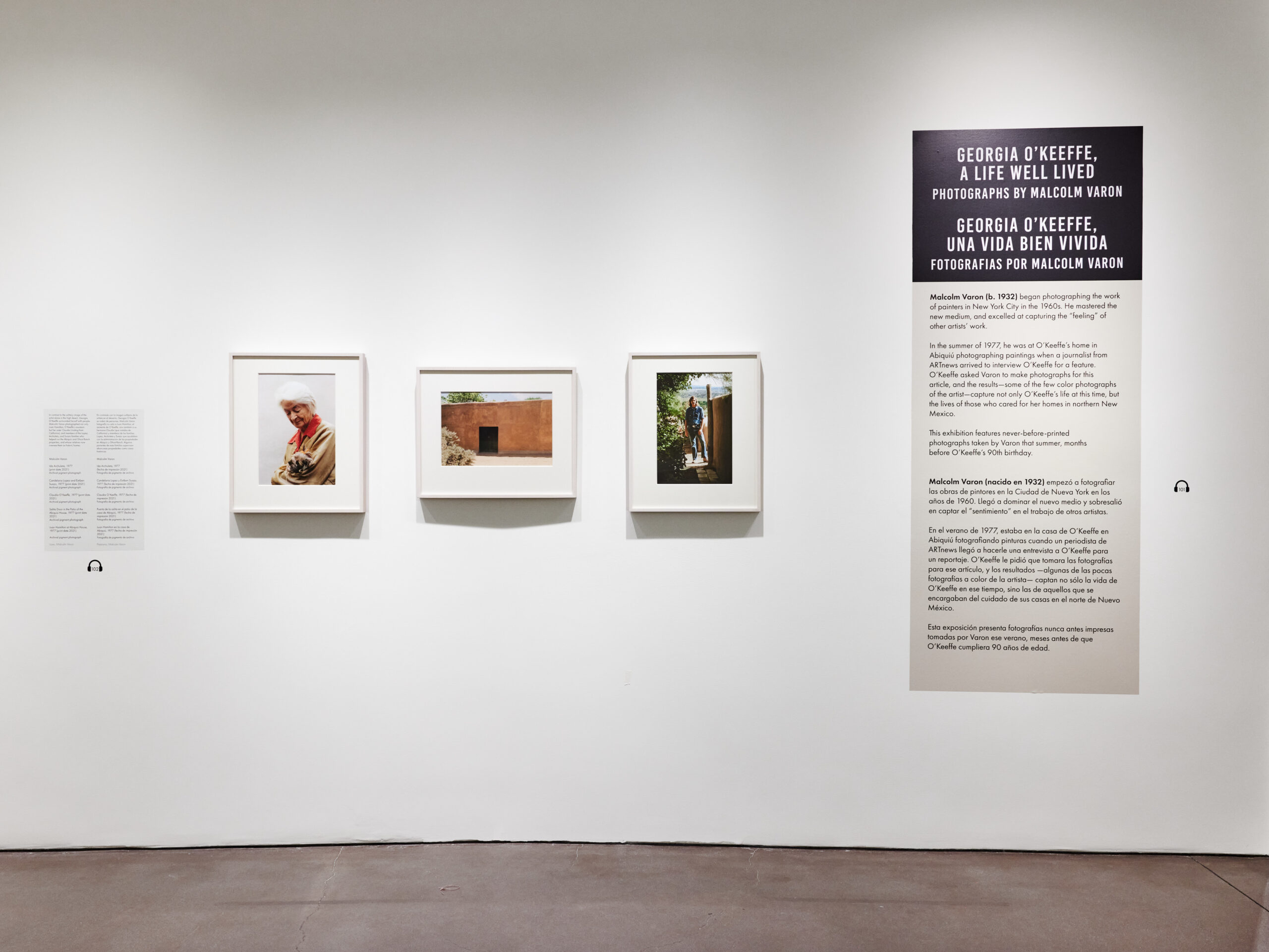 A view of a gallery with white walls and three framed color photographs and a text panel. The room has a dark brown floor.