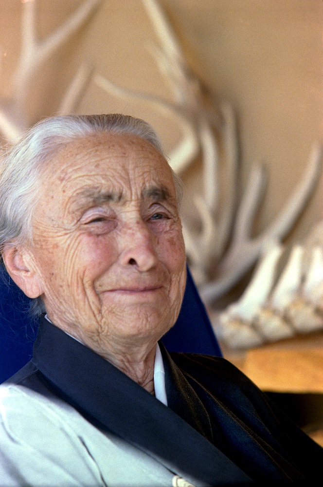 A woman looks straight ahead with a smile and eyes squinting. She has grayish white hair and weathered skin with wrinkles forming around the smile. In the background are large antlers and bones shown against a tan wall.