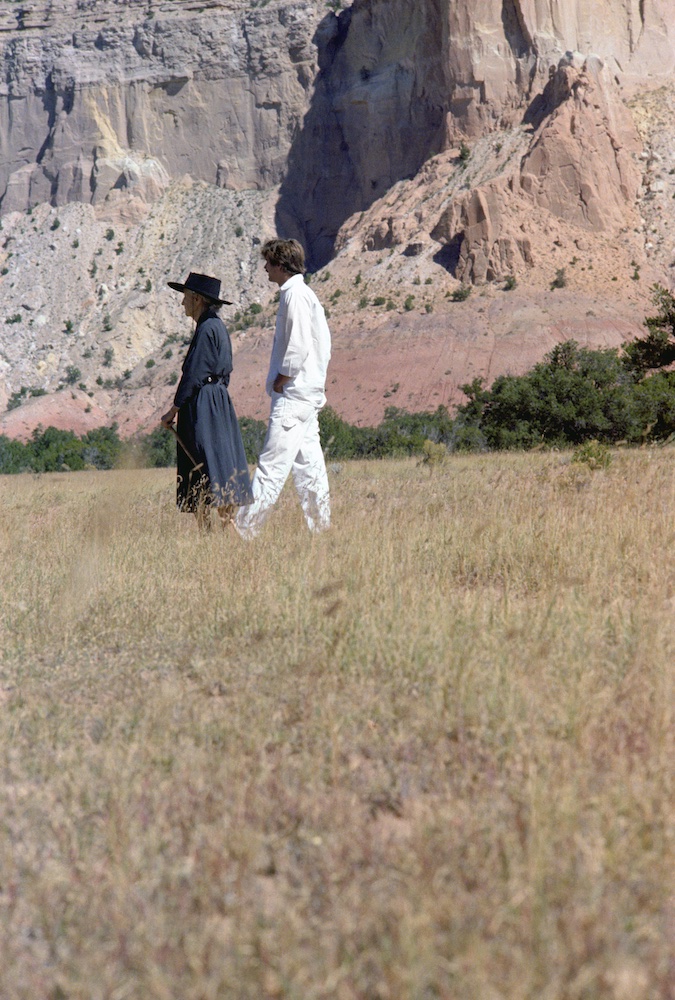 Two people, one dressed in a black flowing dress and the other in a white shirt and pants stroll in the background of a desert meadow with painted rock formations in the background.