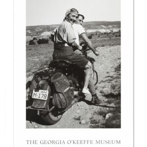 White poster with a black and white photograph in the center and the words 'The Georgia O'Keeffe Museum, Santa Fe, New Mexico' in the bottom. The photograph shows Georgia O'Keeffe sitting behind a companion on a motorcycle. O'Keeffe wear a had cap, goggles, jeans, light shoes and a shirt rolled to her elbows. The motorcycle is on a dirt road and a desert landscape and sky stretches out behind the riders. The motorcycle is turned away and O'Keeffe look back towards the camera. O'Keeffe is smiling.