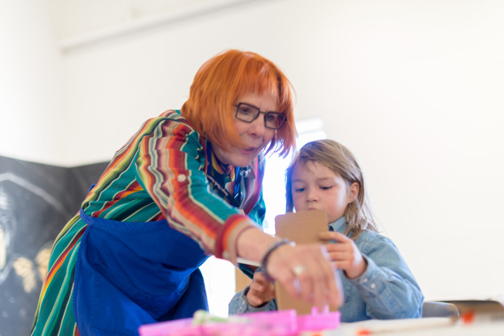 An adult in glasses and a child are bent over a piece of artwork on a table. The adult wears a blue apron.