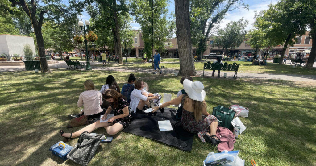 A group of students sit sketching on a green lawn with trees in the background.