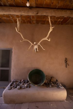 Large antlers attached to a skull hangs on a beige stucco wall. Below it is a tabletop with a selection of stones and a pot on its side. There is part of a door on the left end of the image.