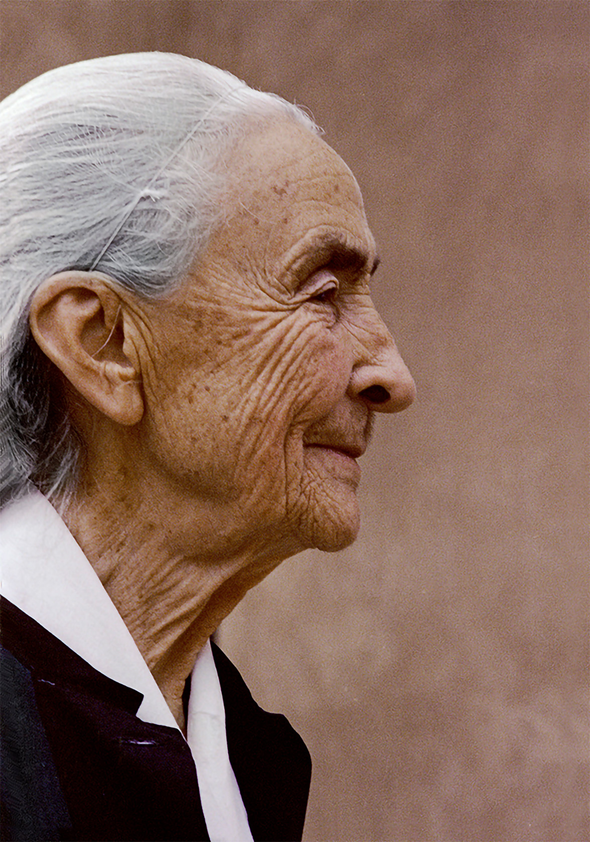 Photograph of a women in profile with white gray hair in a subtle matching hair net. Her skin is peachy-tan and worn with wrikles. She is slightly smiling. The woman wears a black jacket or shirt with a white collar and stands against a tan-colored stucco wall.