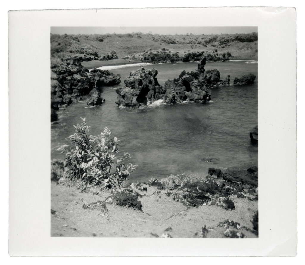 A black and white photograph of Hawaii. In the foreground there is sandy cliff that drops off into the ocean water. In the middle of the water craggy rocks emerge out of the ocean.