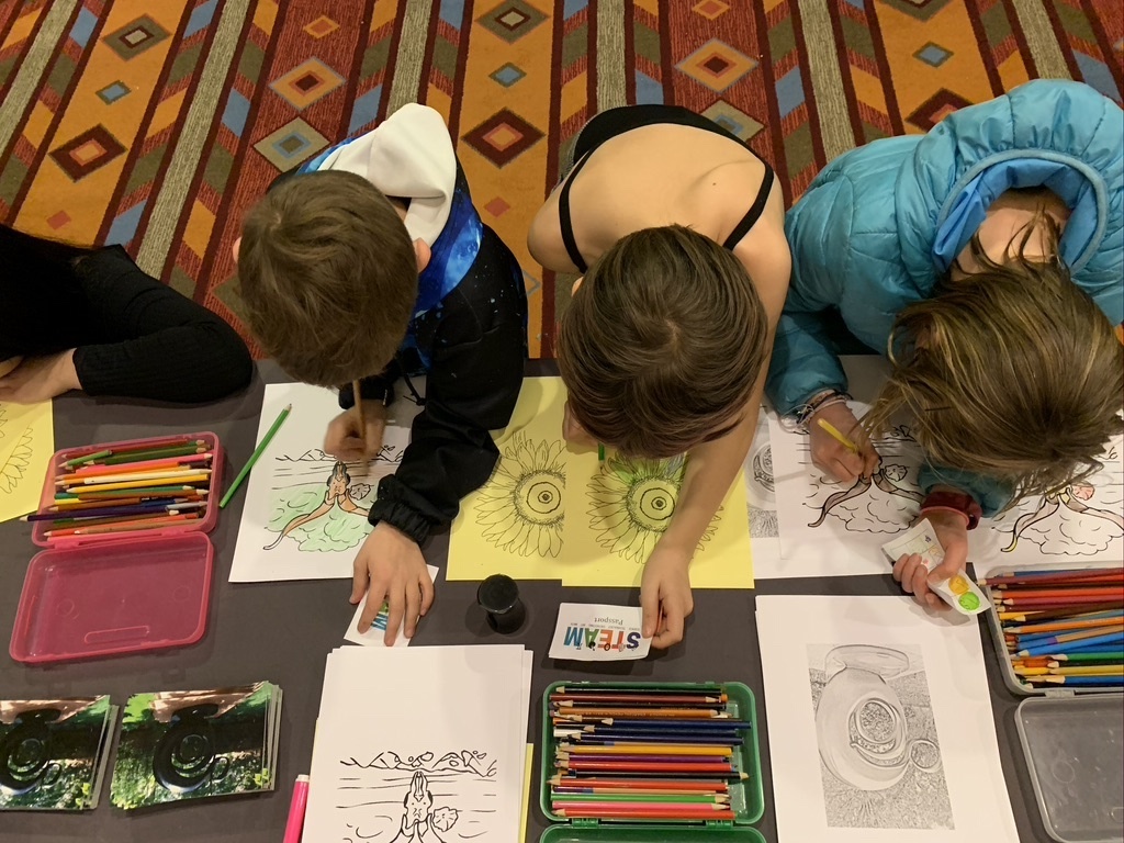 Three children pictured from above working on art pieces on the floor which is covered in art supplies