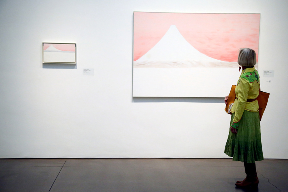 Person dressed in green staring at the large painting 'Untitled (Mt.Fuji), 1960' by Georgia O'Keeffe on a white exhibition wall. Next to the large painting is a small similar depiction O'Keeffe created that same year. Both paintings show a white peak on a peach background.