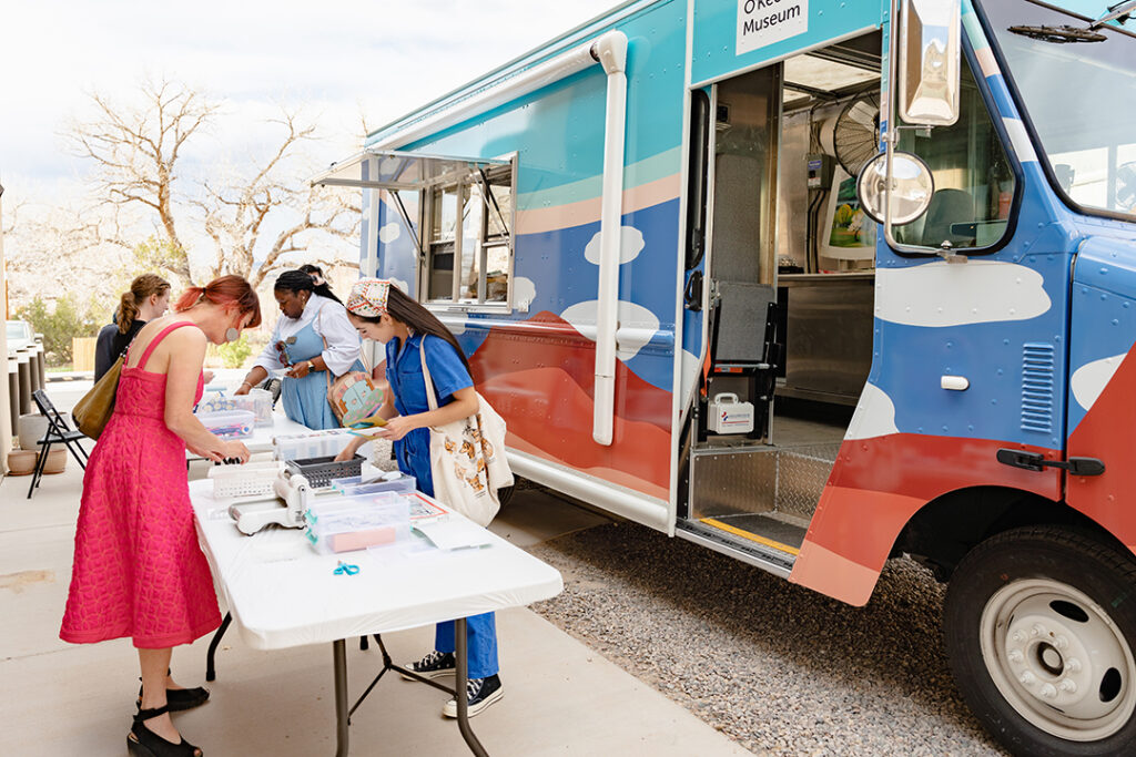 On the right is the Art to G.O. Truck with cloud motifs and an open window facing a few tables. On the left of the frame are tables with art supplies and a group of people standing around them making collages. In the background are trees.