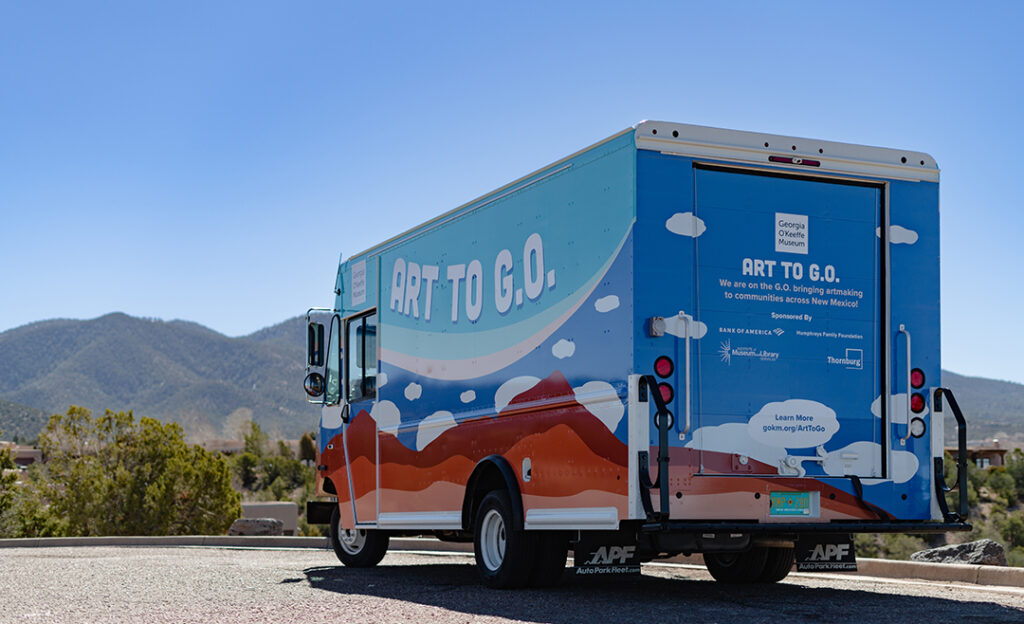 Image of the Museum's mobile creative studio with O'Keeffe-inspired clouds and mountains with the name Art to G.O. on it. The truck faces away from the camera and behind are mountains