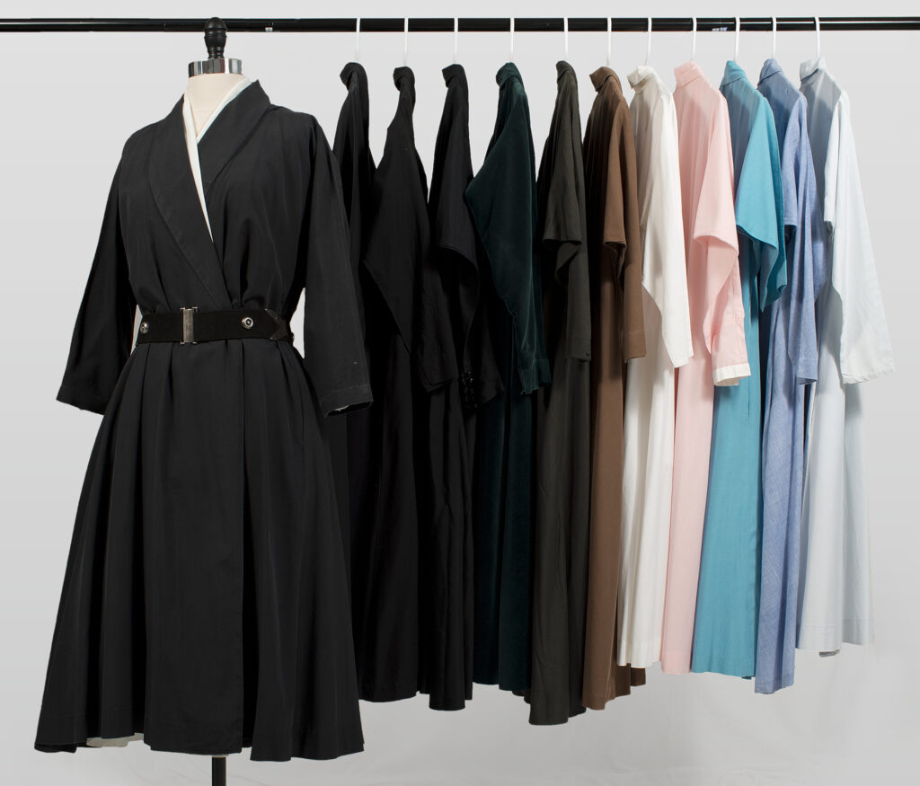 Eleven of O'Keeffe's dresses hang on a rack. Those on the left are dark shades of black and grey, and on the right, they range from white to light blue and pink. A black wrap dress with a belt is on a mannequin apart from the others on the left side of the frame, facing the camera.