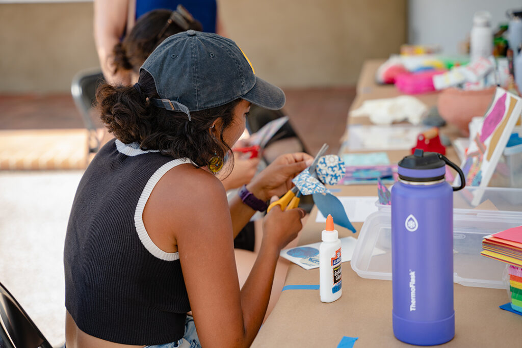 Young person participating in an arts and craft activity. They are wearing a hat and holding some paper and scissors.