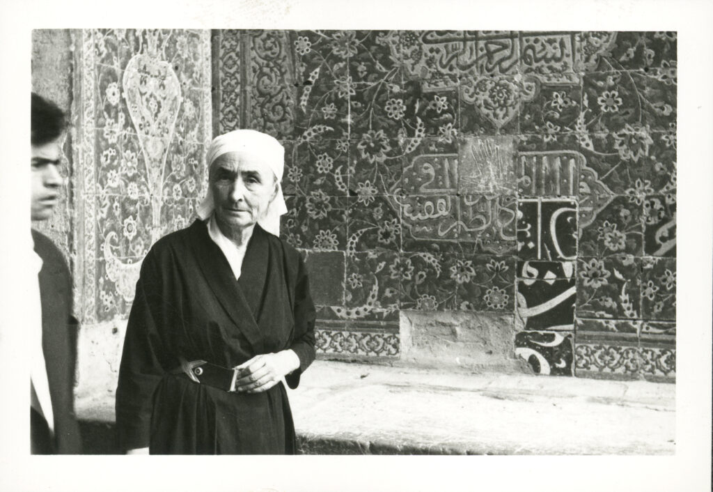 Black and white photograph of Georgia O’Keeffe wearing a dark wrap dress and a light scarf around her head at a Mosque. Behind her are the intricate tiles of the Mosque wall and to the left of the frame is gentleman passing through in front of the camera.