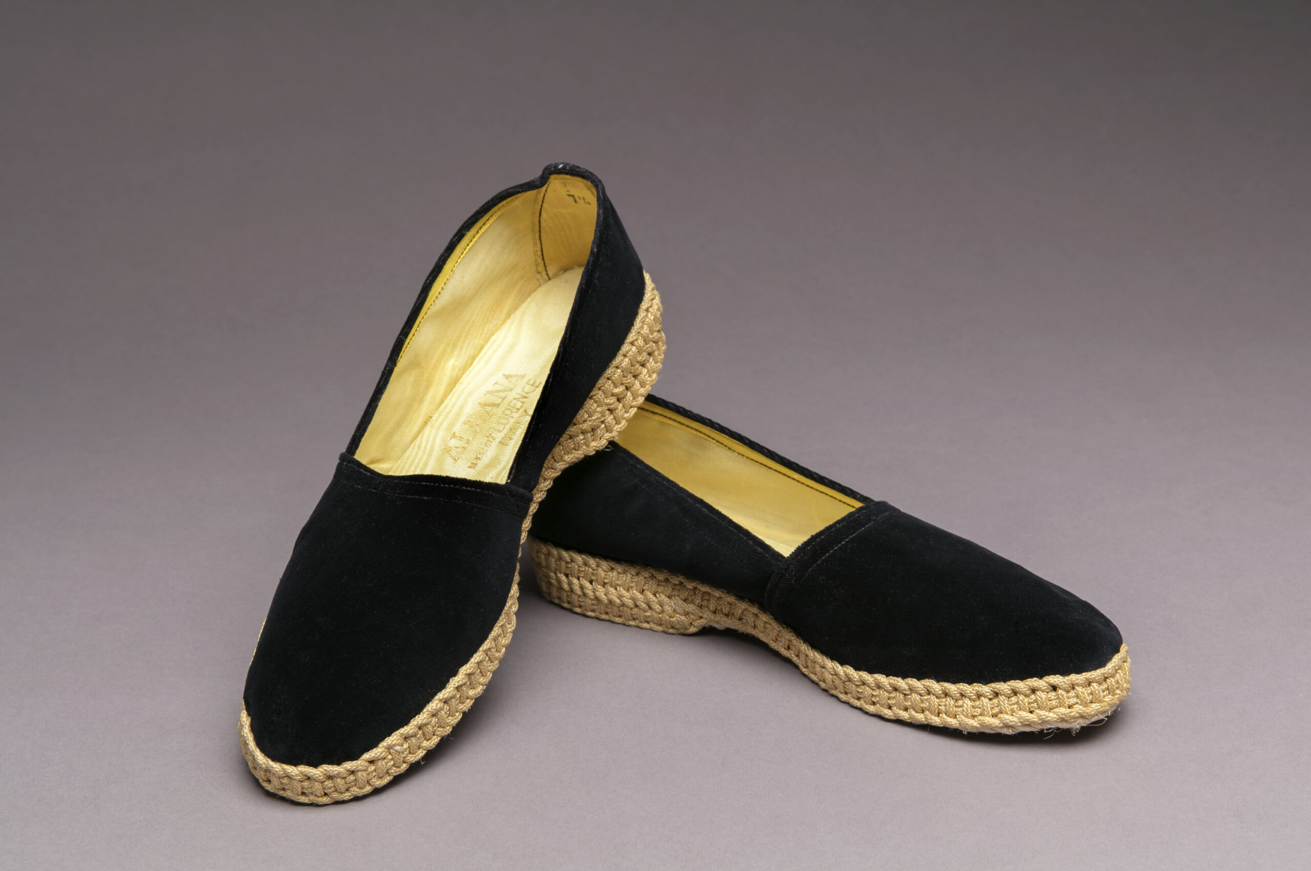 A photograph of black velvet espadrille shoes posed on top of each other. The heel and border of the bottom of the sure is made of beautifully woven rope. The shoes are lined with yellow silk.