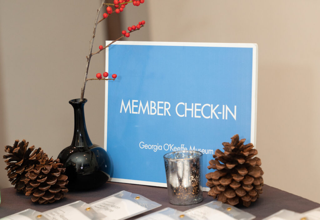 Photograph of a blue 'Member Check-In' sign on a table surrounded with candles, pinecones, and branches in a vase. On the bottom of the frame are name tags