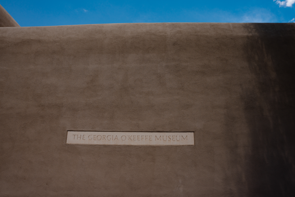 Adobe wall exterior of the Georgia O'Keeffe Museum with a sign of the Museum's name. Above the wall is a sliver of blue sky and a little cloud