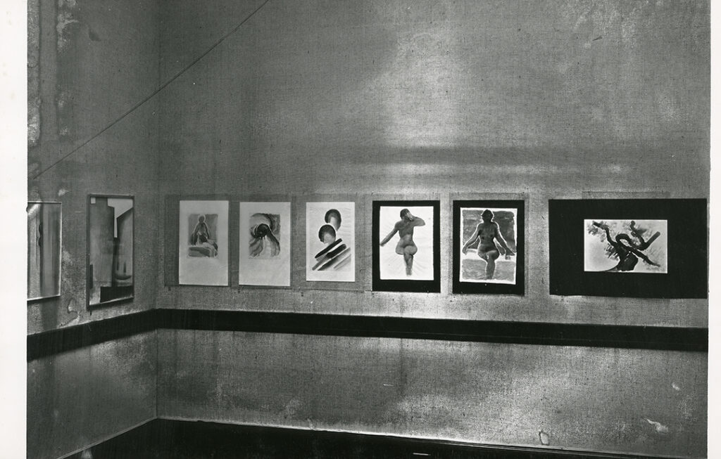 Black and white photograph showing several of O'Keeffe's watercolors on display. On the wall in focus are eight of O'Keeffe's watercolors, including a wall of an exhibition with three nude watercolors and several more. Under the hanging paintings is a dark moulding.