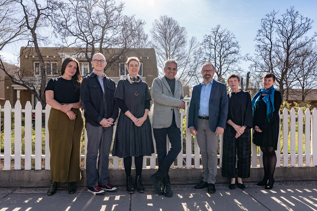 Group of seven individuals standing in front of a white wooden fence looking towards the camera and smiling. Behind them are bare trees against a blue sky and the very top of a building.