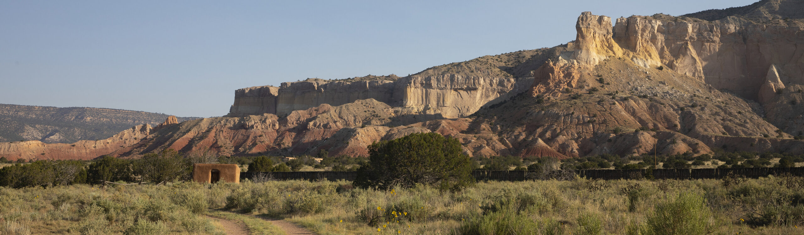 Landscape photograph of tall cliffs in shades of golden and red rock with a blue sky in the background. Green bushes and trees are at the bottom of the photo and to the left is a dirt road and a small square adobe building with a door.
