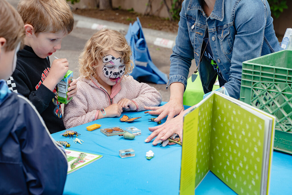 Photograph of three children, one in face paint, leaning over a table looking intently at an adult's hands as they demonstrate an art activity.
