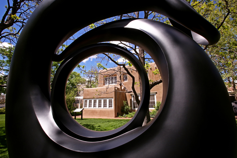 Exterior of the Museum's Research Center and Library viewed though a large dark cast of O'Keeffe's sculpture 'Abstraction.' The adobe building, green grass and blue sky are visible through the sculpture's circular shape