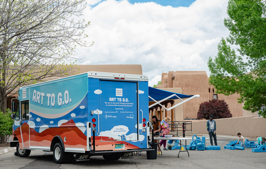 Photograph of the 'Art to G.O.' truck with cloud motifs on the side in a parking lot under a tree. To the right of the truck and under it's awning, people are gathered over a table for art activities. Blue foam blocks with children playing in them take up the right side of the frame and behind the truck are adobe buildings and large white clouds.