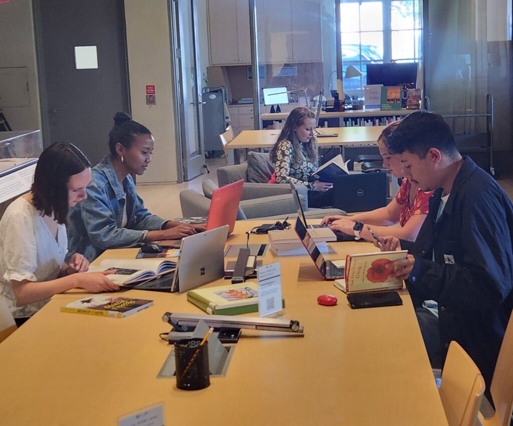 5 people sitting at a table working on laptops and referencing books.