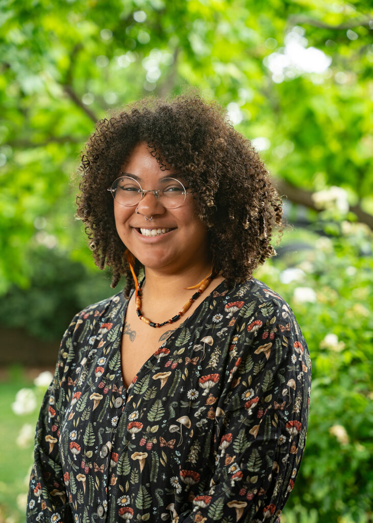 Photograph of a person with glasses and a necklace smiling at the camera in front of greenery.